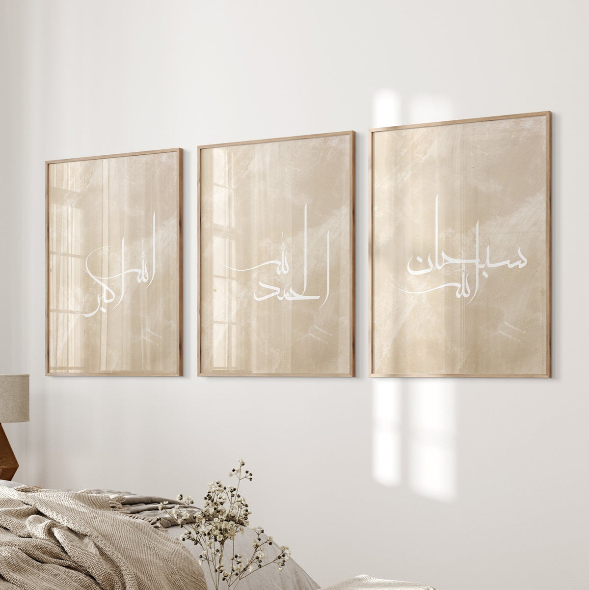 A set of three beige and white Islamic wall art prints with Arabic calligraphy, displayed above a bedside table in a room.