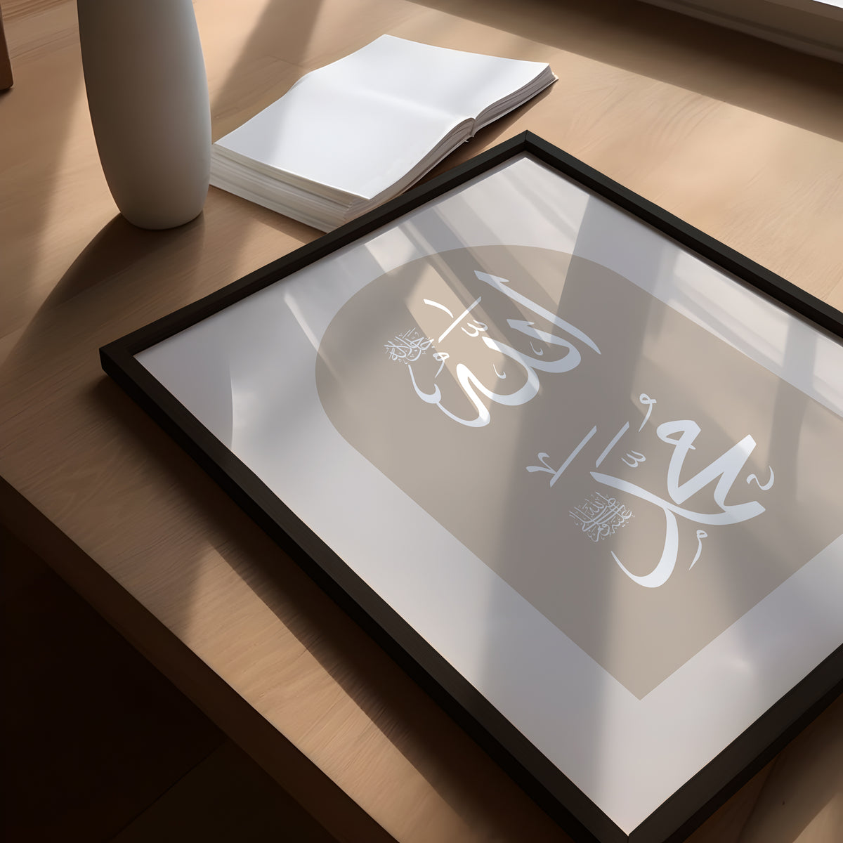 A set of three beige-framed Islamic wall art pieces is placed on a office desk, featuring Arabic calligraphy that reads "Hayya alasalah" and "Haya alal falah," which translates to "Come to prayer" and "Come to success," respectively, along with the names of Allah and Prophet Muhammad saw.