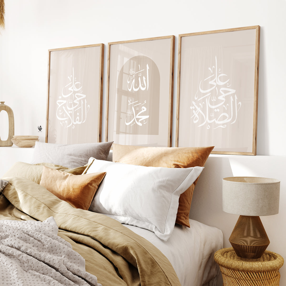 A set of three beige-framed Islamic wall art pieces is placed above a bed with pillows, featuring Arabic calligraphy that reads "Hayya alasalah" and "Haya alal falah," which translates to "Come to prayer" and "Come to success," respectively, along with the names of Allah and Prophet Muhammad saw.