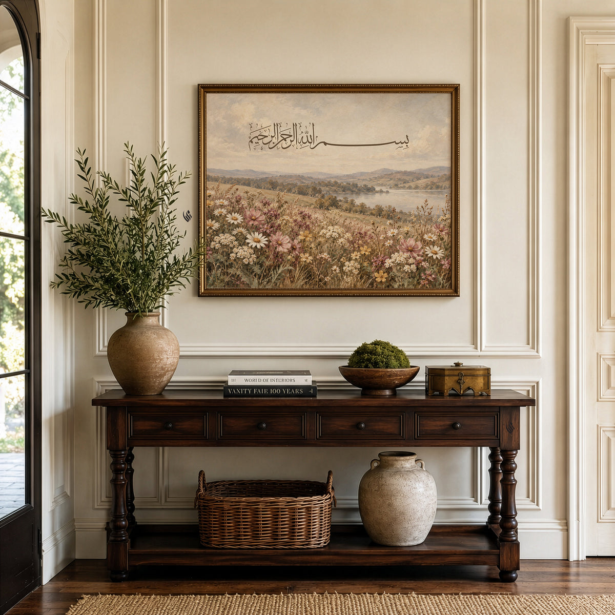 Bismillah Arabic calligraphy wall art in an ornate gold frame, displayed above a traditional dark wooden console table in a neutral hallway.
The landscape artwork features a peaceful lake, rolling countryside hills and a foreground filled with pink, cream and white wildflowers.
Styled with a large ceramic vase of olive branches, decorative books and rustic accessories against elegant cream panelled walls.