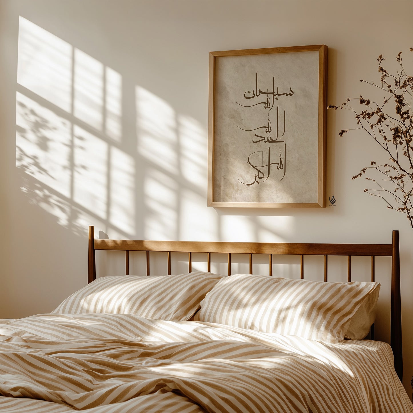 A styled bedroom scene with a wooden bed frame, cream and tan striped bedding, and a framed Arabic calligraphy print on the wall. Sunlight with window shadow patterns is cast across the wall and bedding.