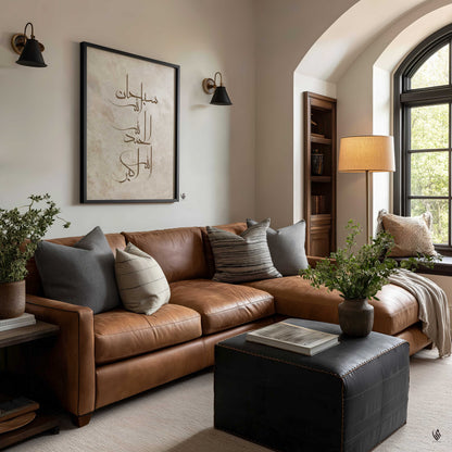 A styled living room featuring a brown leather sectional sofa with gray and striped accent pillows, a black leather ottoman, a window, and a framed Arabic calligraphy print on the wall.