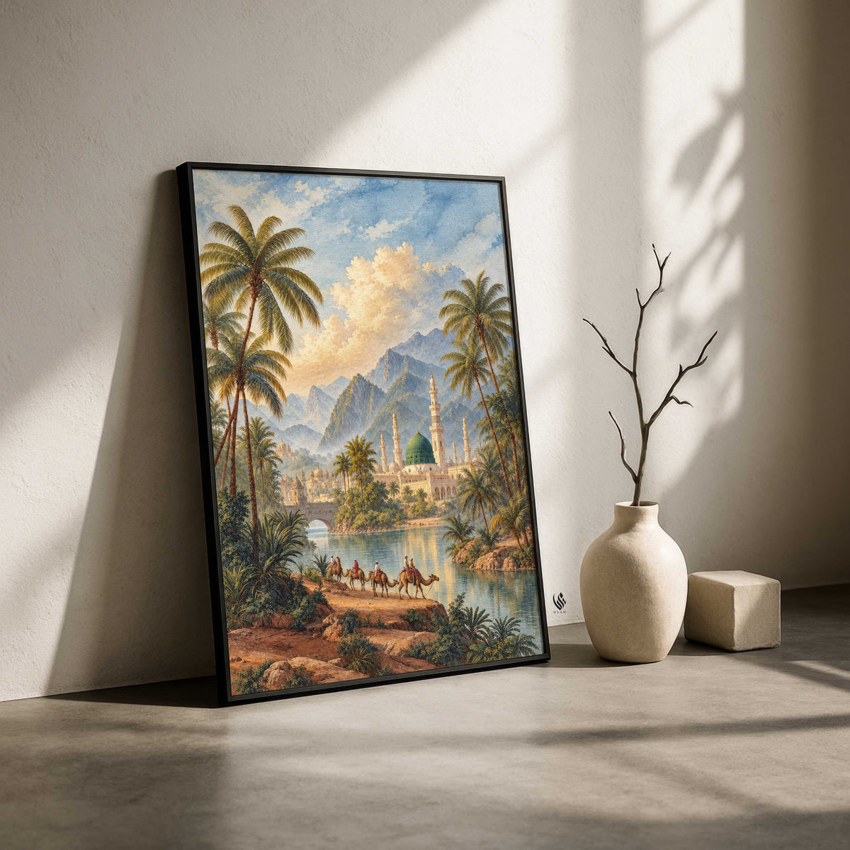 A framed landscape art print leaning against a wall on a concrete floor, next to a ceramic vase with a bare branch and a small block. The artwork depicts a tropical oasis with palm trees, mountains, a mosque, camels, and a river under a blue sky.