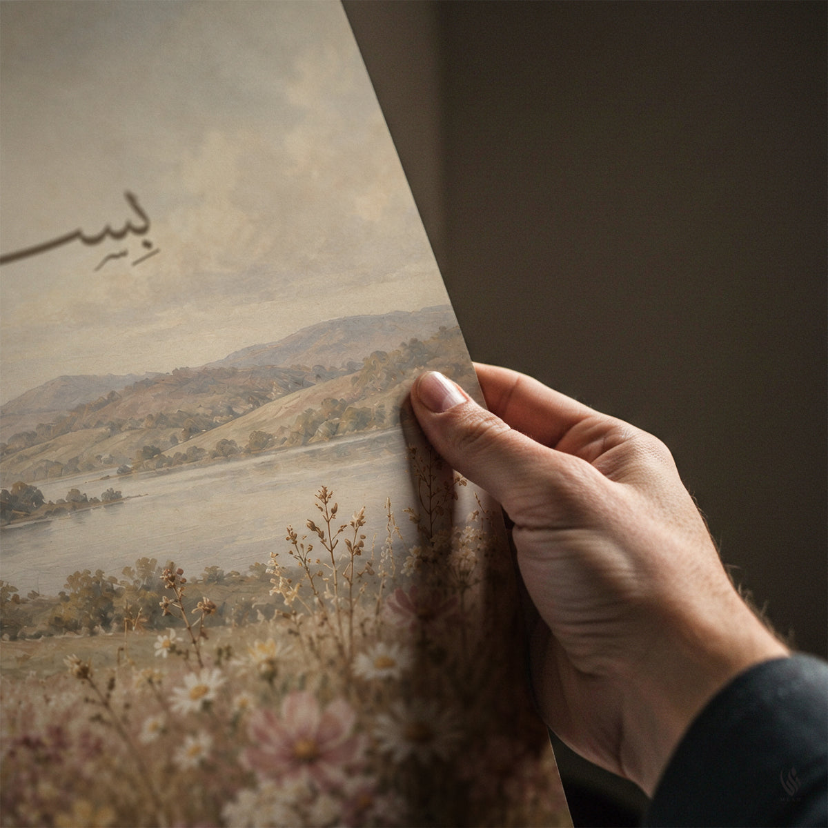 A hand holds a vintage-style art print or poster showing a pastoral landscape with hills, a river, and wildflowers. The print is partially folded over, revealing a dark brown background behind it.