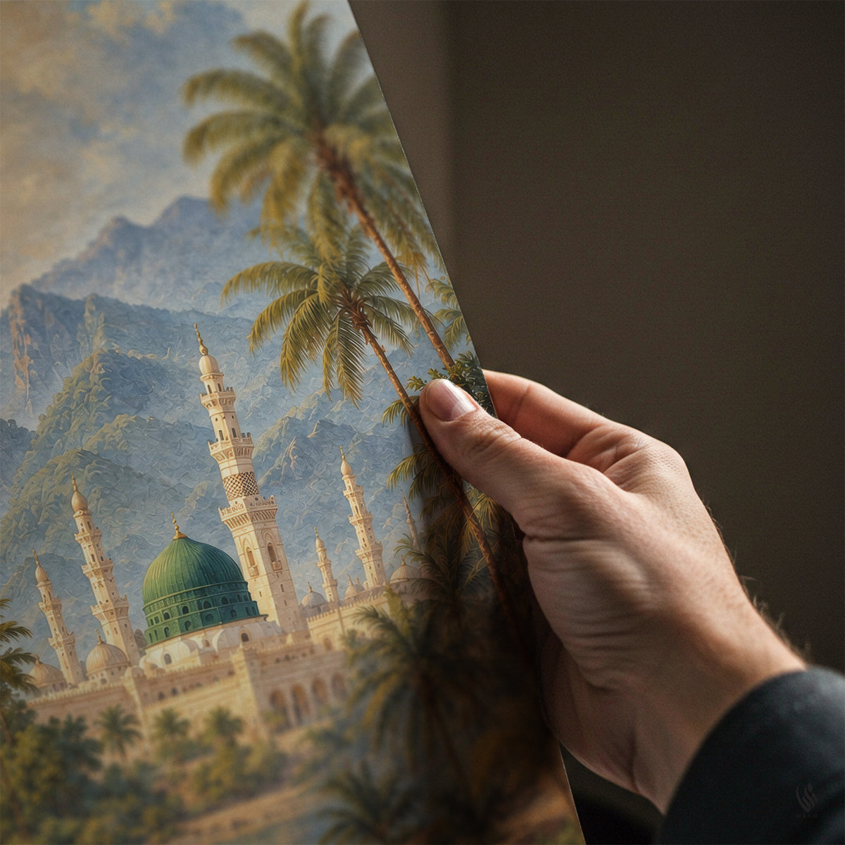 A hand holds a large art print or poster showing a scenic painting of a mosque with green domes, palm trees, and mountains, against a dark brown background.