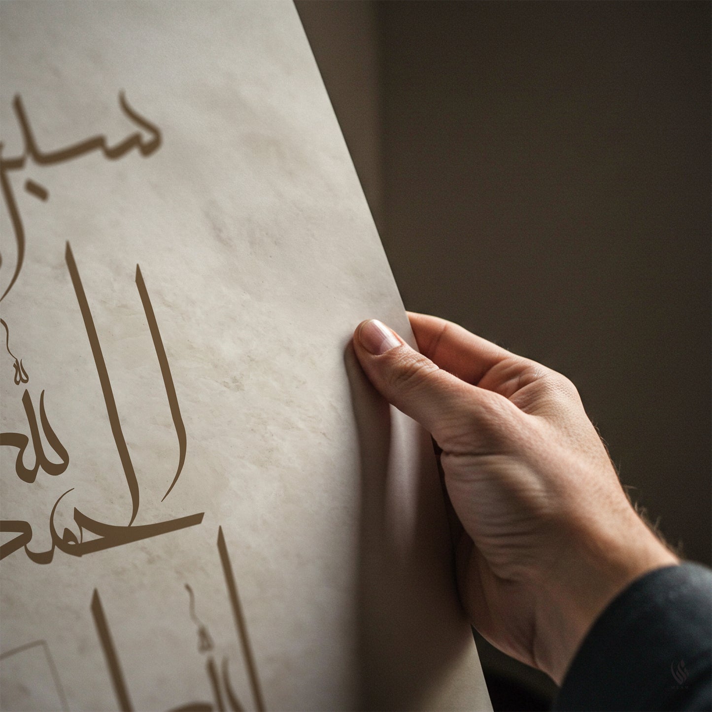 A close-up of a hand holding a large sheet of textured beige paper with elegant Arabic calligraphy printed on it, set against a dark, neutral background.
