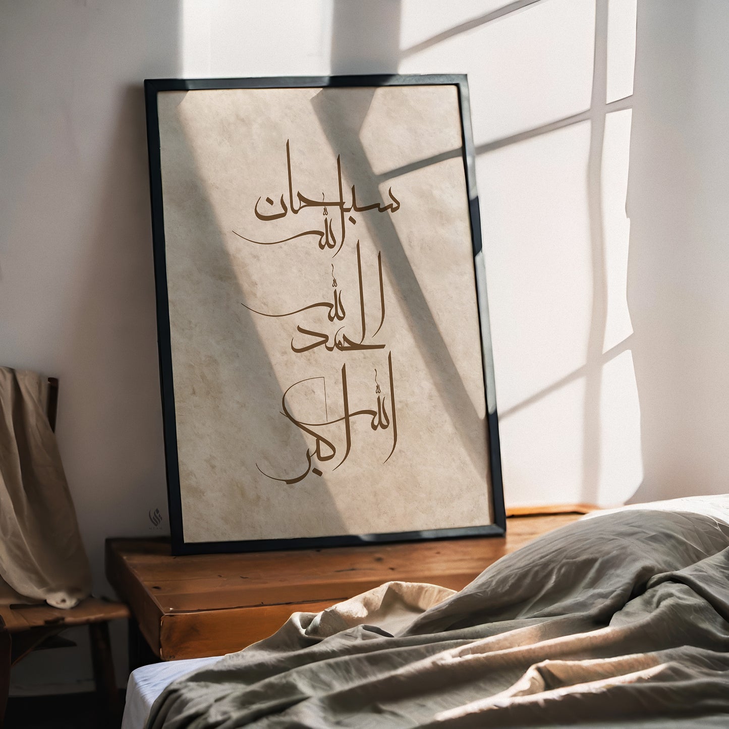 A framed Arabic calligraphy art print leaning against a wall on a wooden table next to a bed with rumpled linens, lit by warm natural sunlight with window pane shadows.