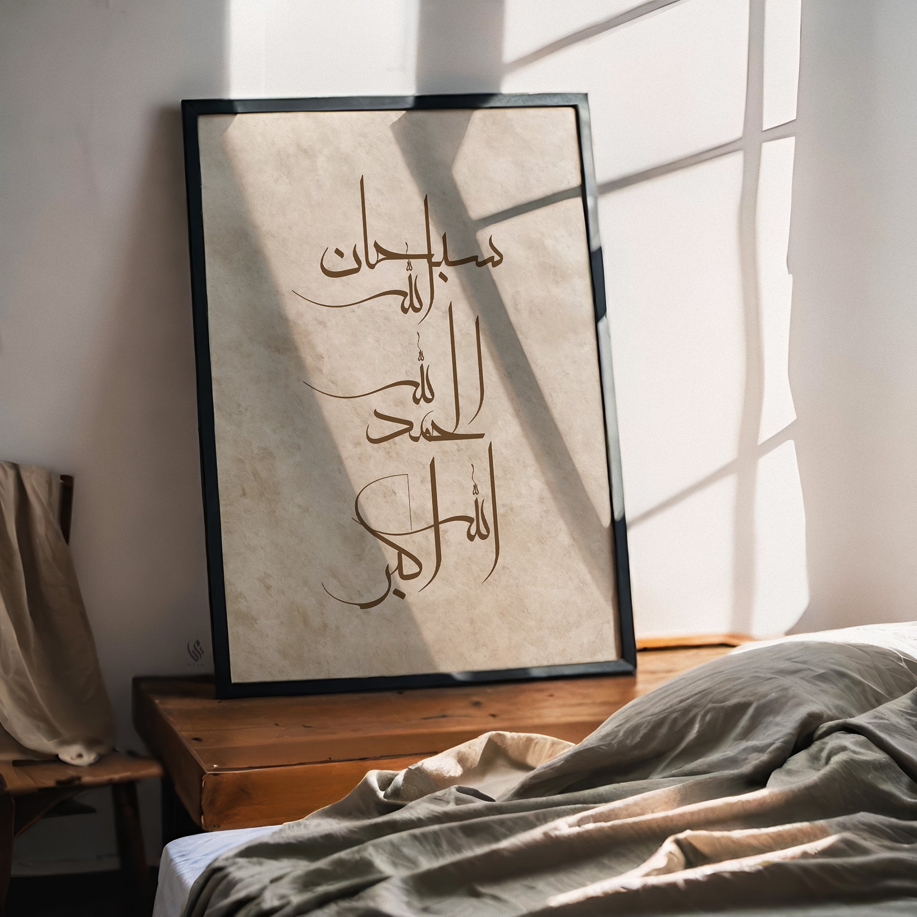 A framed Arabic calligraphy art print leaning against a wall on a wooden table next to a bed with rumpled linens, lit by warm natural sunlight with window pane shadows.