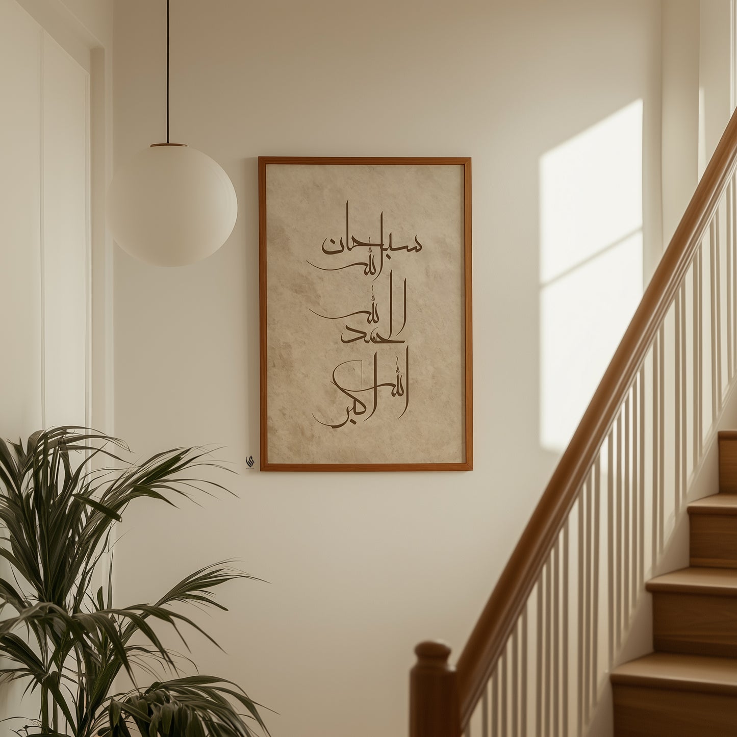 Framed Arabic calligraphy wall art hanging on a cream-colored wall in a modern interior setting with a wooden staircase, a white globe pendant light, and a potted plant.