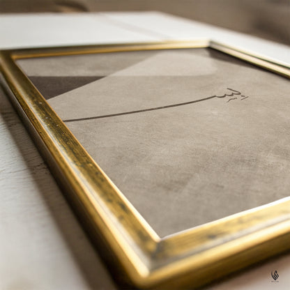 A close-up of a framed art print featuring minimalist Arabic calligraphy in black on a textured beige background, enclosed in a gold-toned frame.
