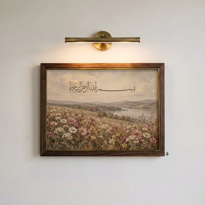Vintage Bismillah Arabic calligraphy print displayed in a dark wooden picture frame with gold inner detailing beneath a brass wall light.
The Islamic landscape artwork features fine brown Arabic lettering above a peaceful countryside lake, rolling hills and a colourful wildflower meadow.
Soft white daisies, dusty pink blossoms and muted golden grasses create a delicate botanical composition against a warm, lightly textured sky.