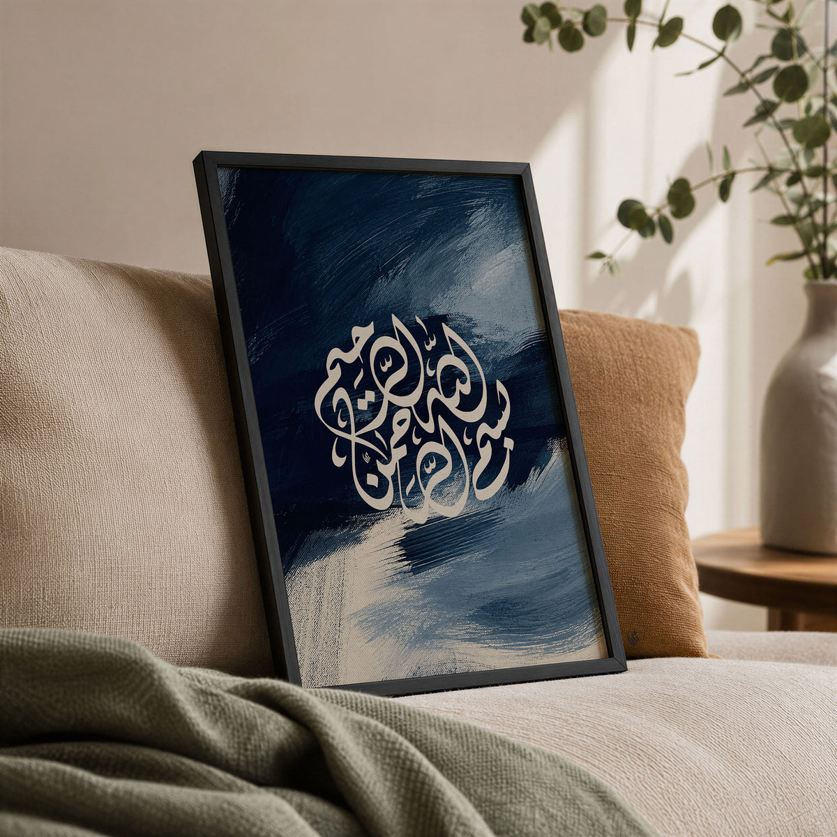 Framed Bismillah Islamic art leaning on a beige sofa, with cream Arabic calligraphy, layered blue brushwork, warm cushions and greenery.