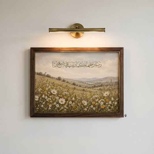 A framed landscape art print featuring a field of white and yellow wildflowers under a cloudy sky with Arabic calligraphy, mounted on a white wall below a brass wall sconce.
