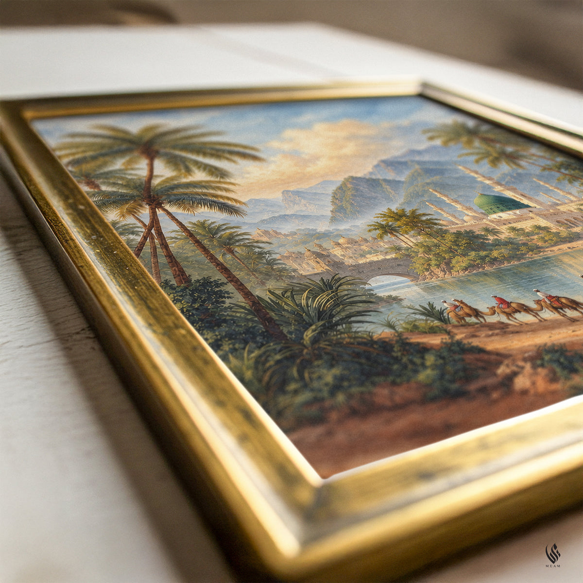A framed vintage landscape painting featuring palm trees, mountains, and camels, set in an ornate gold frame on a light wooden surface.