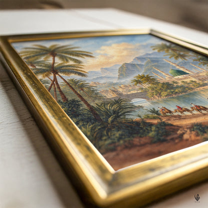 A framed vintage landscape painting featuring palm trees, mountains, and camels, set in an ornate gold frame on a light wooden surface.