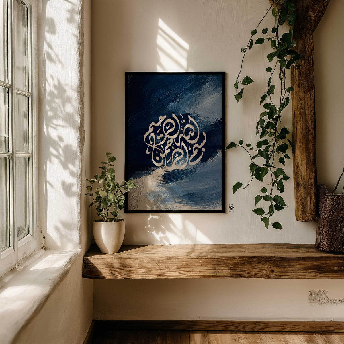 Navy blue Bismillah Arabic calligraphy print in a black frame above a rustic wooden bench, surrounded by leafy plants and window light.