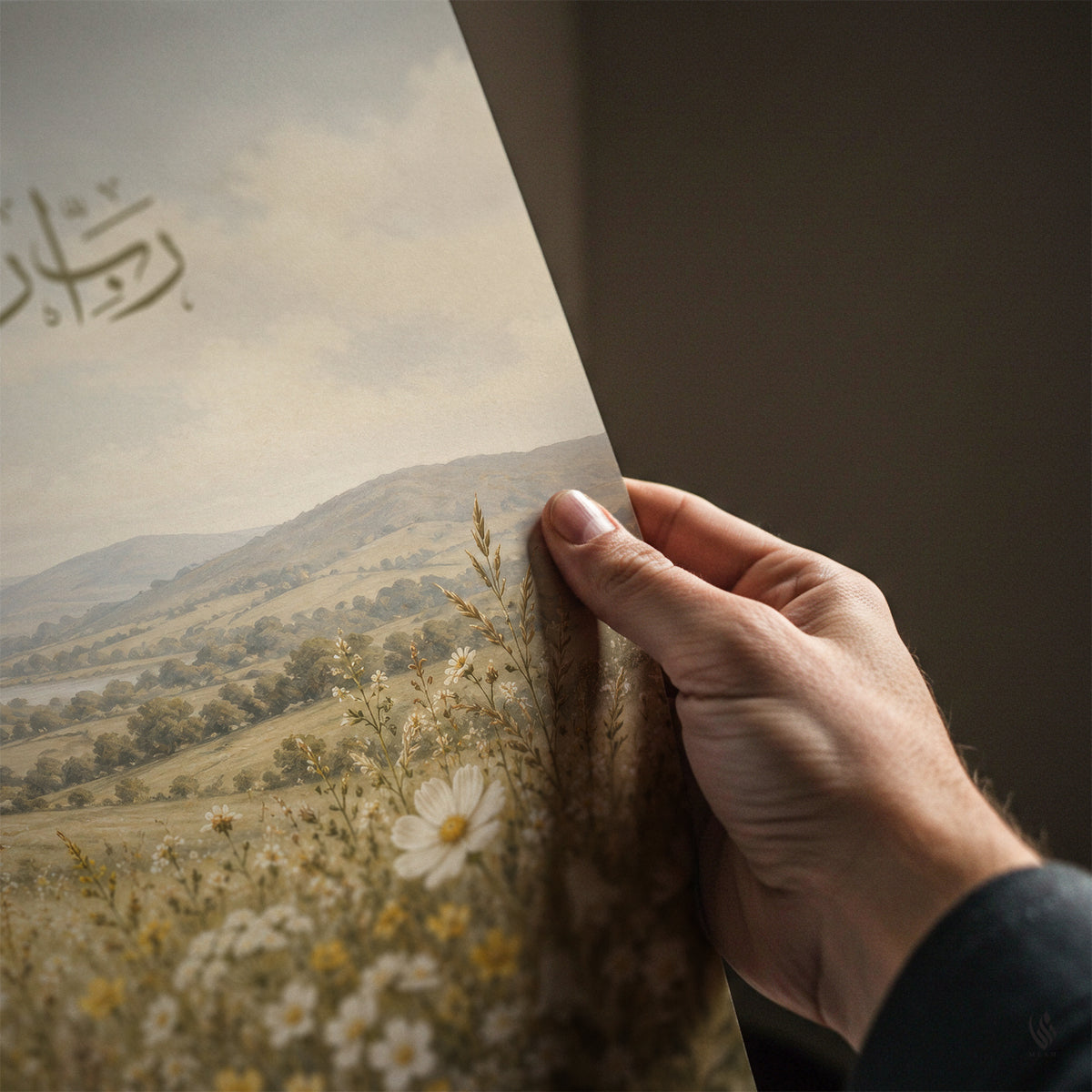 Close-up of a hand holding the edge of an unframed Rabbi Irhamhuma art print, revealing muted mountain scenery, meadow blossoms and part of the Quranic text.