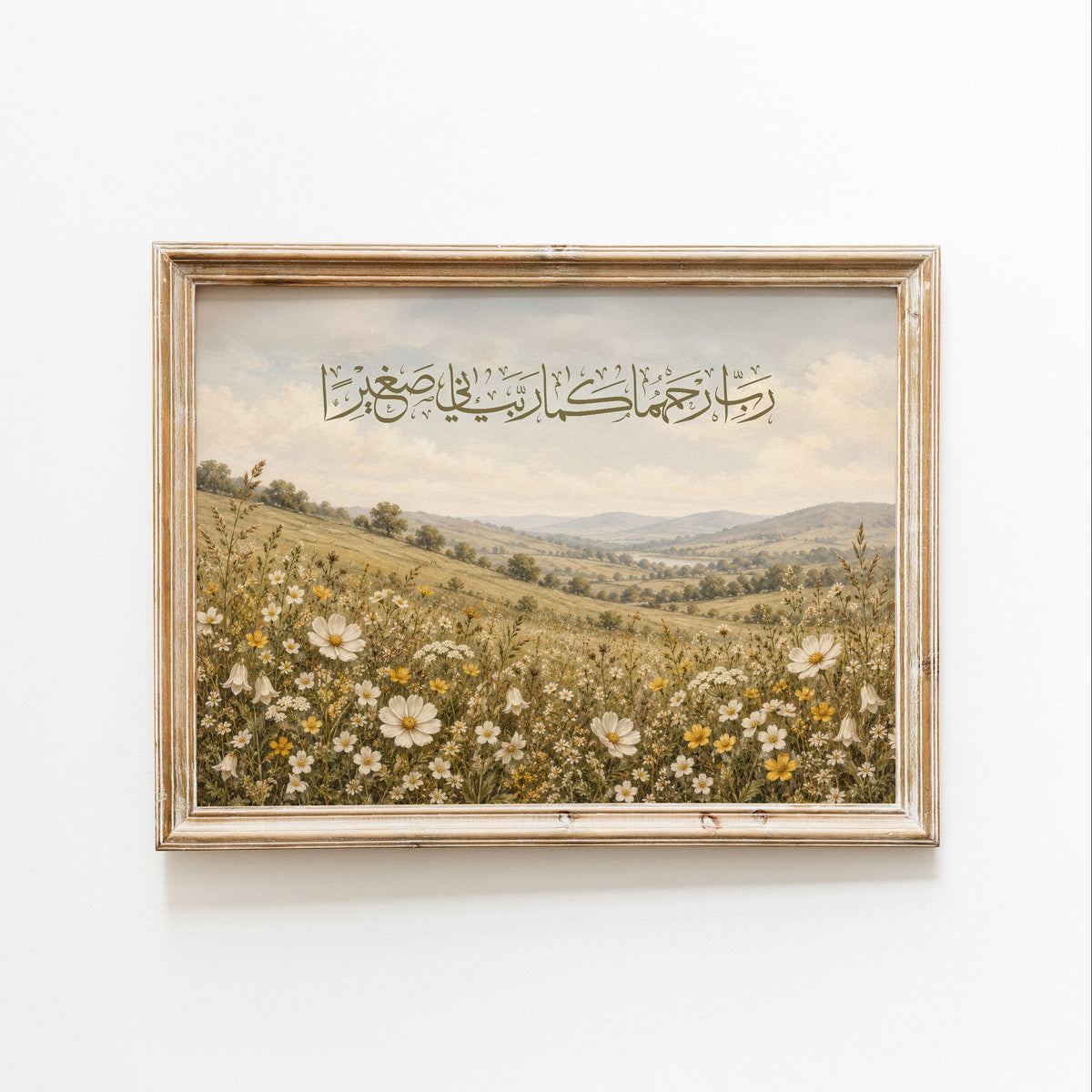 A framed art print depicting a scenic landscape of rolling hills covered in white and yellow wildflowers under a cloudy sky, with Arabic calligraphy written across the top. The print is housed in a light-colored wooden frame and mounted on a plain white wall.