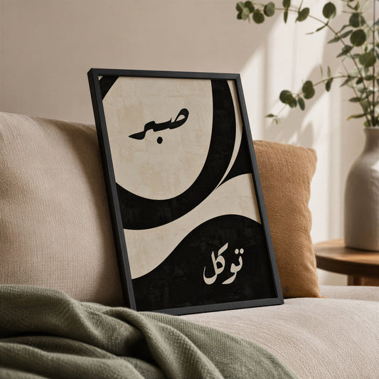 Sabr and Tawakkul Arabic calligraphy print in a black frame leaning on a beige sofa, with black and cream abstract artwork, green throw and leafy plant.
