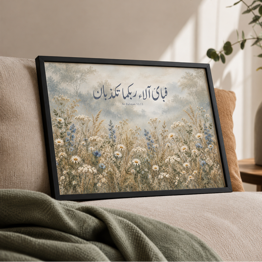 Landscape Islamic wall art in a black frame featuring Surah Ar-Rahman 55:13 in Arabic calligraphy above a misty wildflower meadow with white daisies, delicate blue flowers and soft trees. Displayed on a beige sofa with a sage green throw, tan cushion, eucalyptus vase and warm natural sunlight.