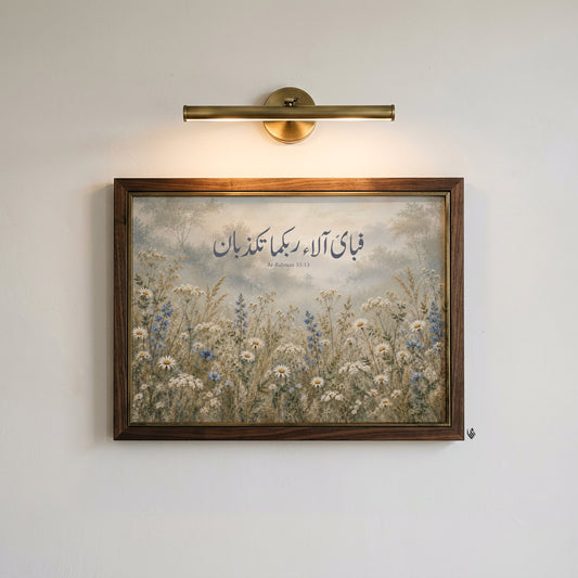 Surah Ar-Rahman 55:13 Arabic calligraphy wall art in a dark wood frame beneath a brass picture light, set against a plain cream wall with muted botanical colours.